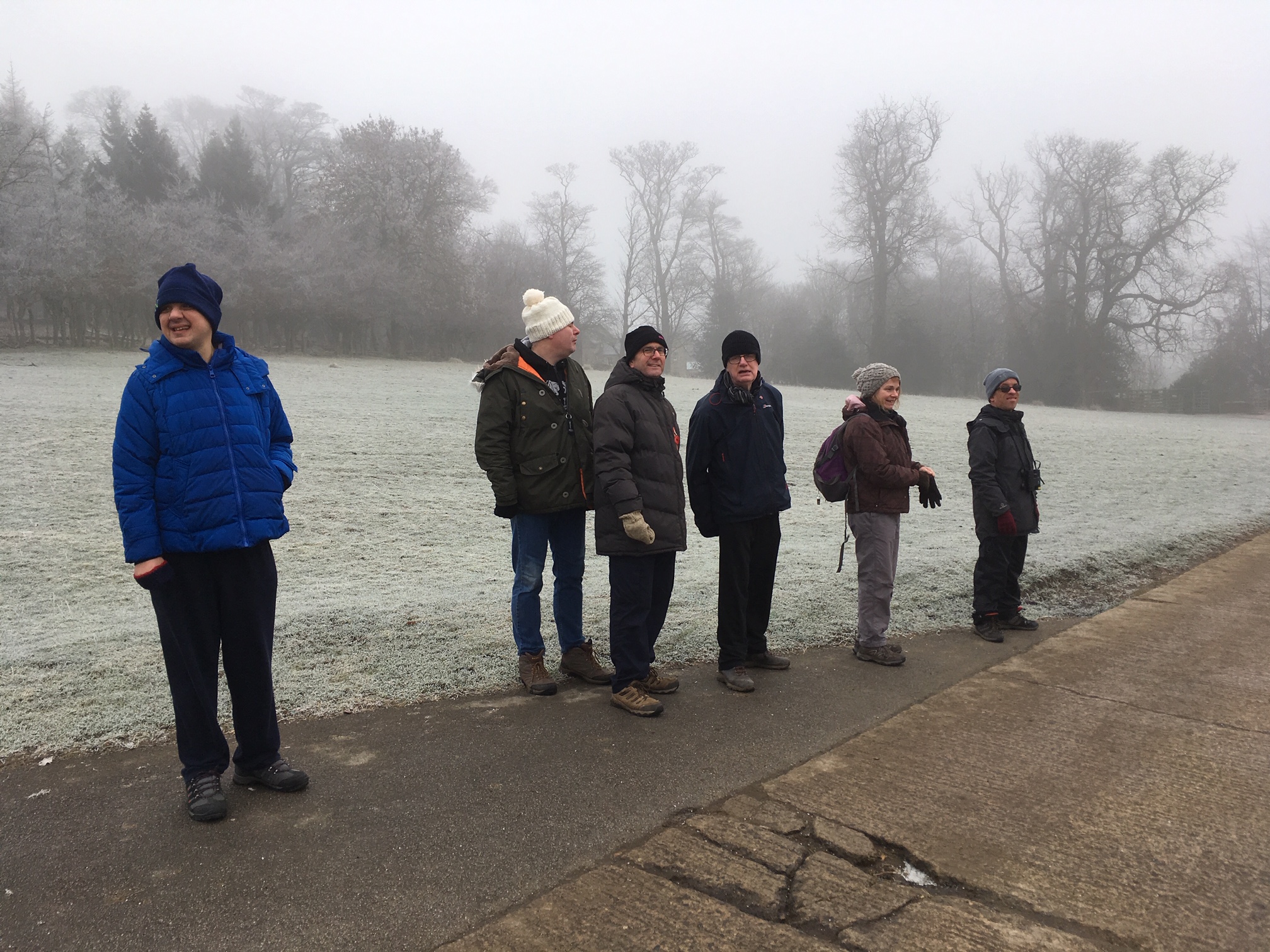 Six people with woolly hats on walking on a track on a cold, foggy day with grass and trees in the background.