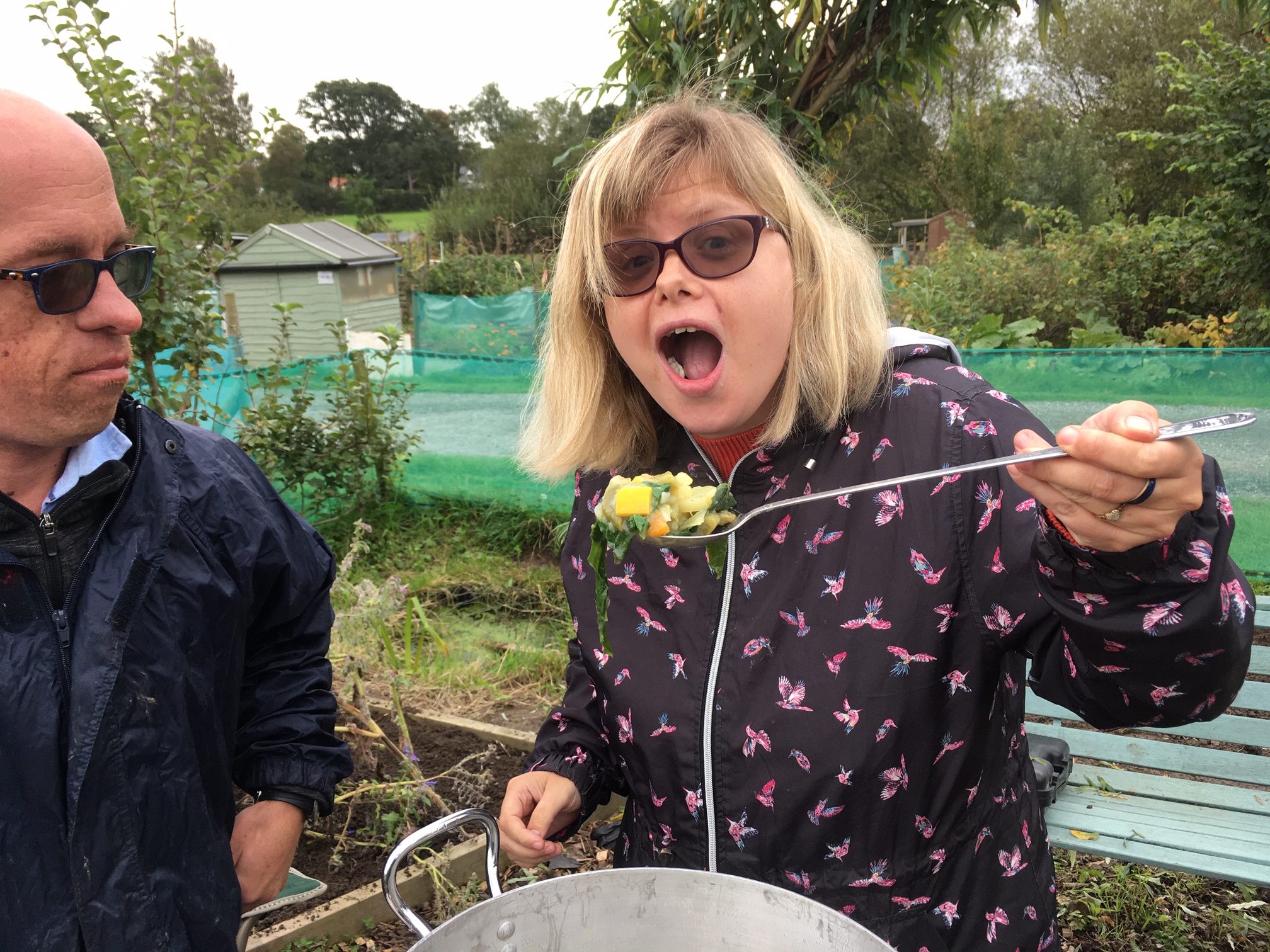 A woman at an allotment eating a big spoonfull of vegetable stew.