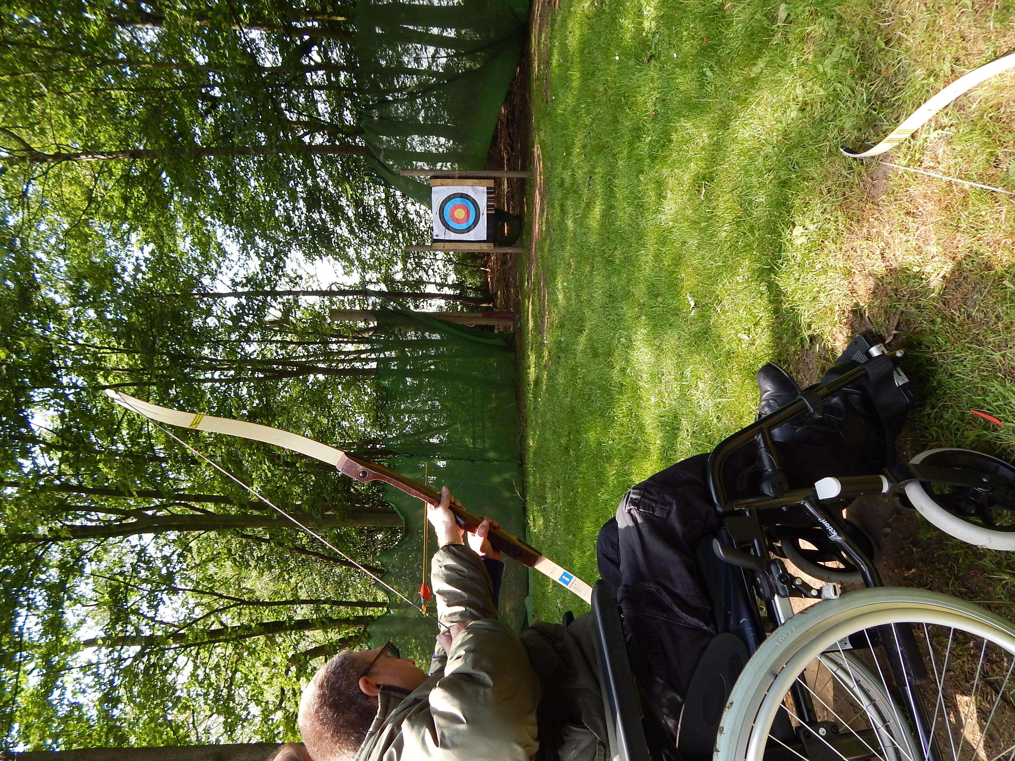 A man in a wheelchair doing archery on grass with woods behind.