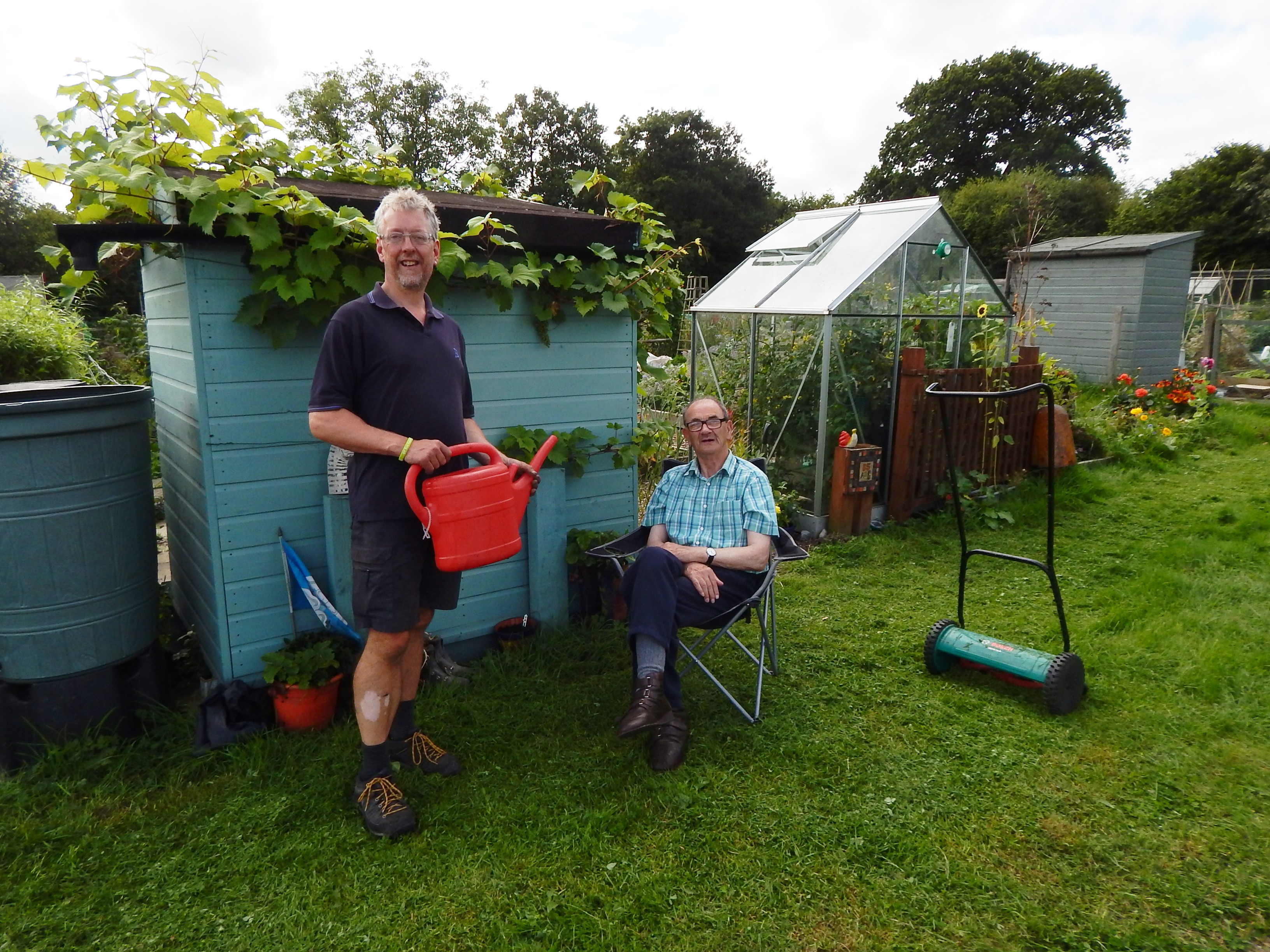 Two men at an allotment by a shed; one is sitting down, the other is standing holding a watering can. 