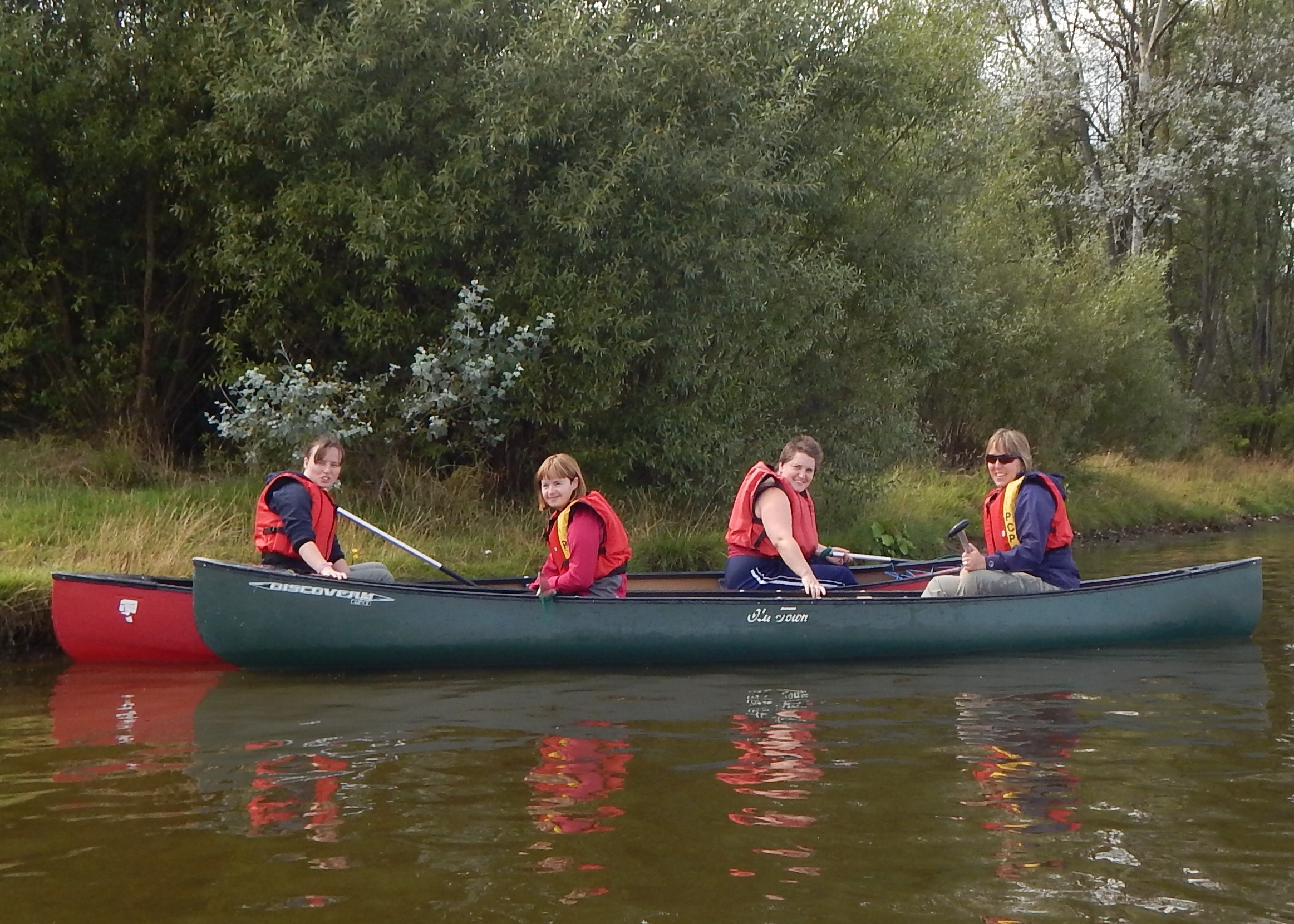 Four women in a canoe on the water with trees behind