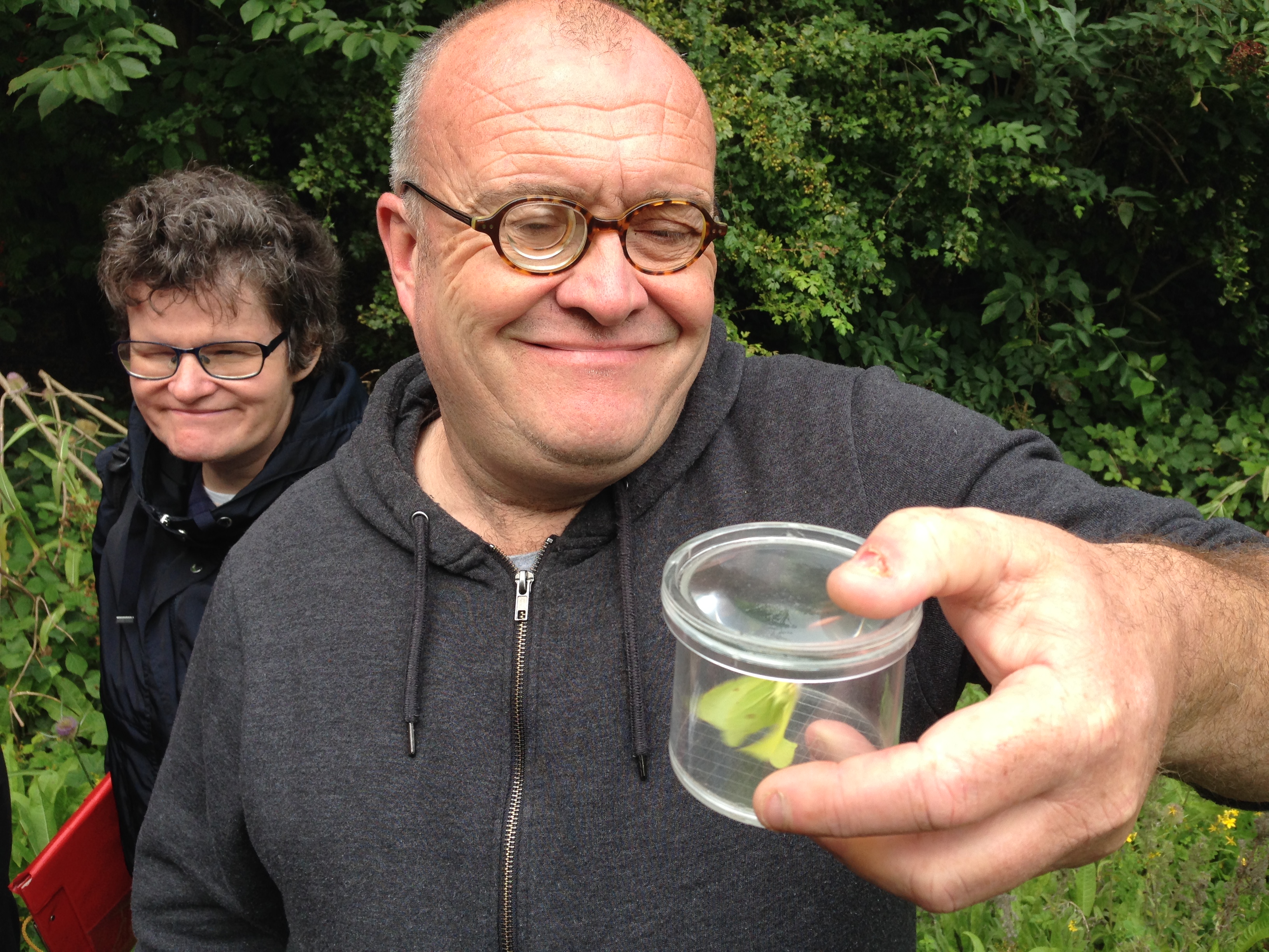 A man smiling and holding a clear container with a butterfly in. A woman is standing behind looking.