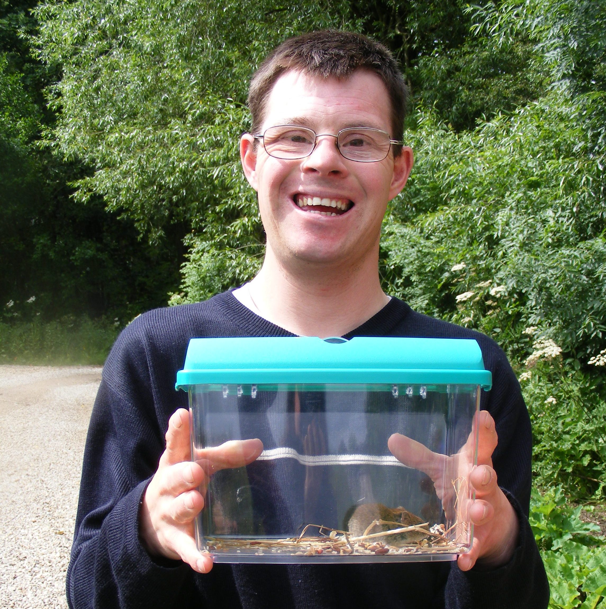 A man smiling with trees behind. He is holding a small container with a small bank vole in.