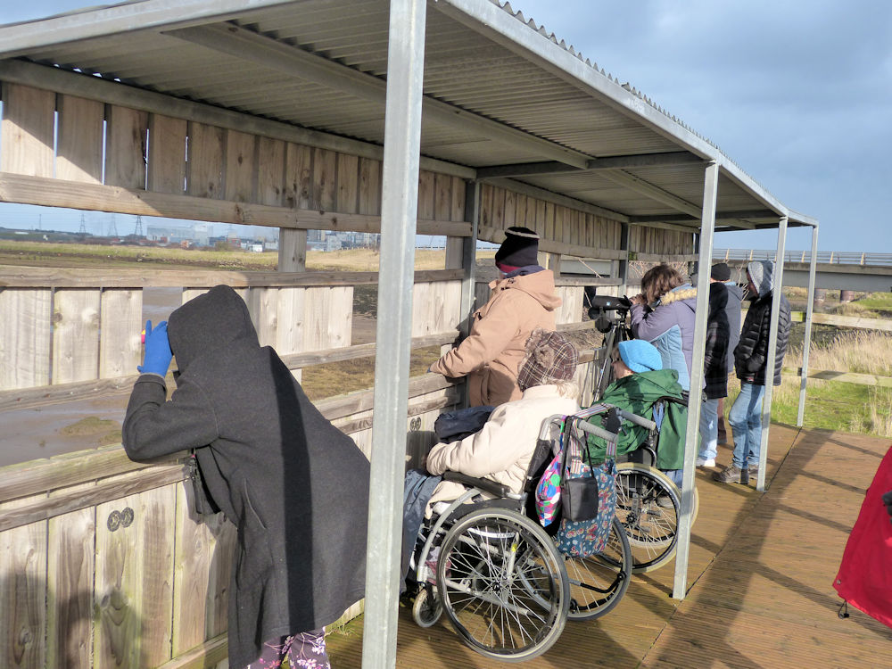 A group of people, some in wheelchairs looking out of a wildlife hide across water.