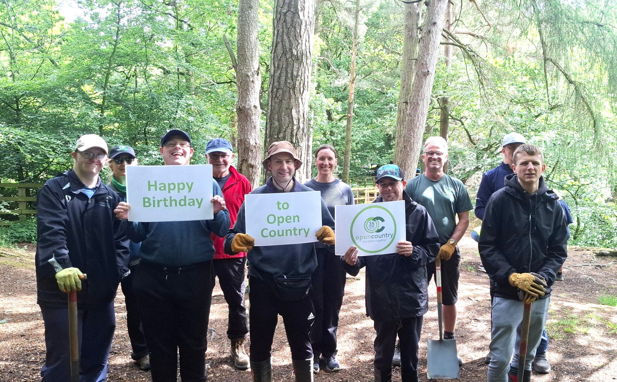 A group of people in a wood hold up signs wishing Open Country a happy birthday