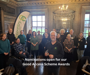 A group of people in an ornate wood panneleed room. At the back is an Open Country flag with green and white logo. At the front is a man, our chief officer David, holding a glass trophy in the form of a thumbs up sign. Text on the photo reads: Nominations open for our Good Access Scheme awards.