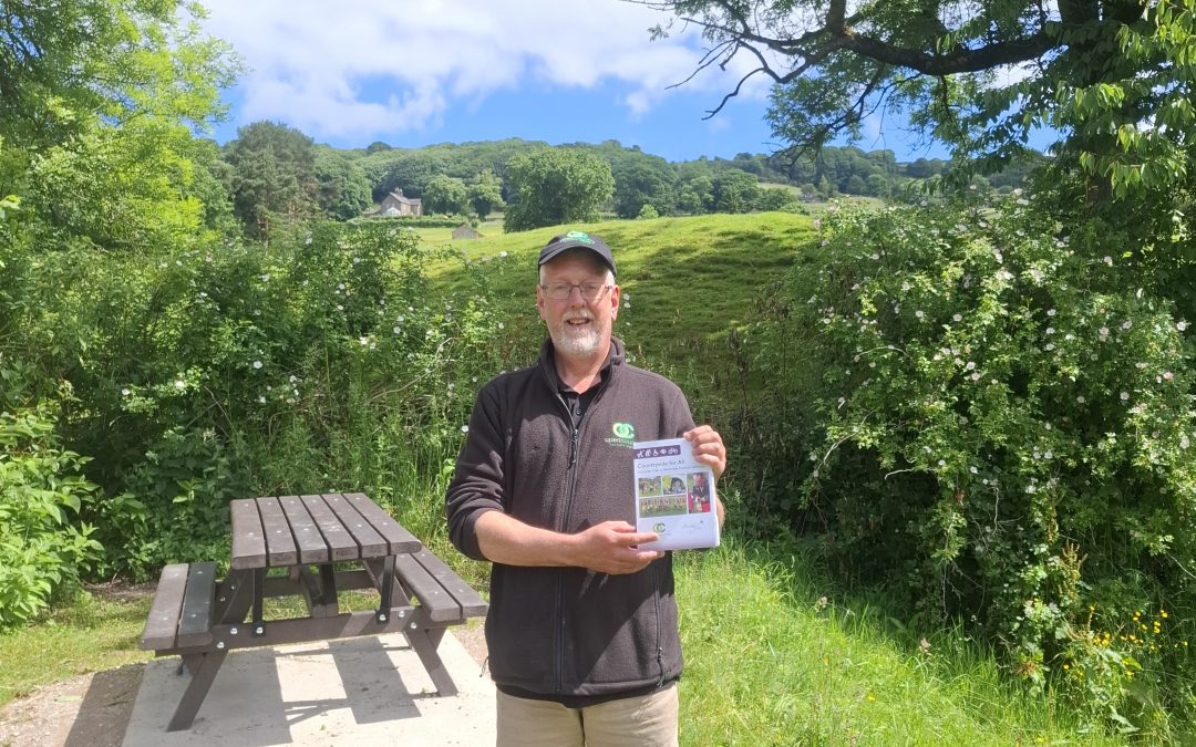 A man stands in a pretty picnic area. He is holding a booklet detailing accessible walks in Nidderdale
